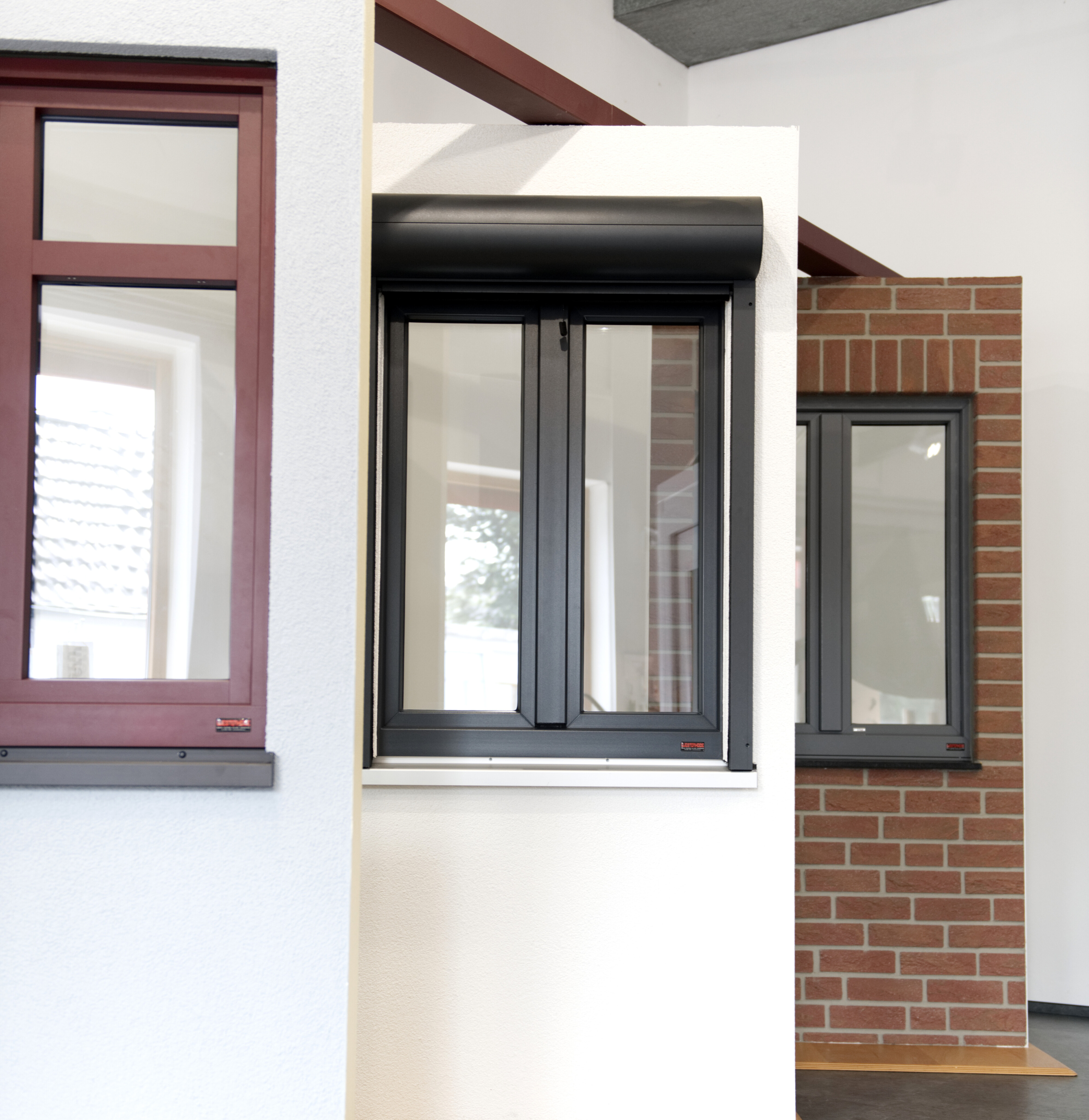 Gruppenbild von drei Fenster-Elementen in verschiedenen Wandtypen. In einem Fenster ist eine Vorbau-Aluminium-Rollade eingebaut mit rundem Kasten in dunkelgrau.